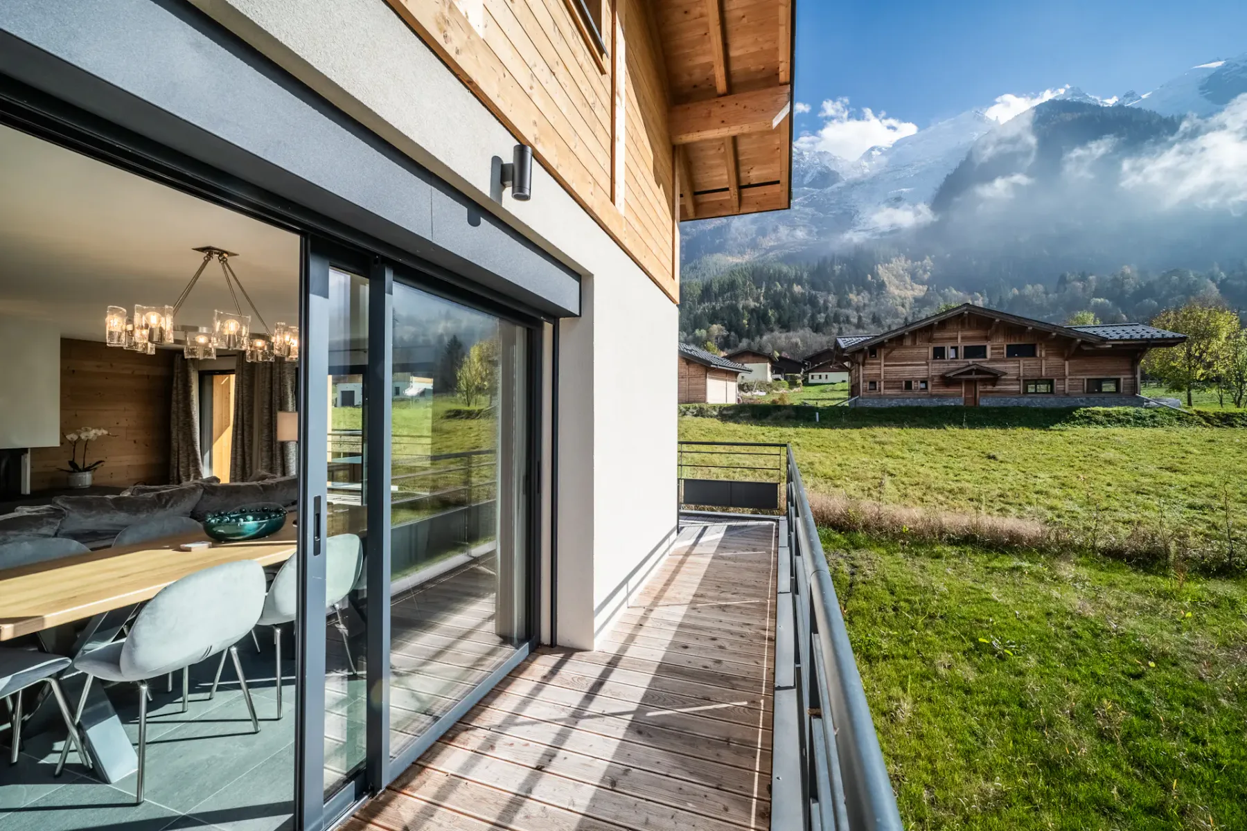 Una terraza de madera junto a una casa moderna da a un campo verde y un pueblo de montaña.