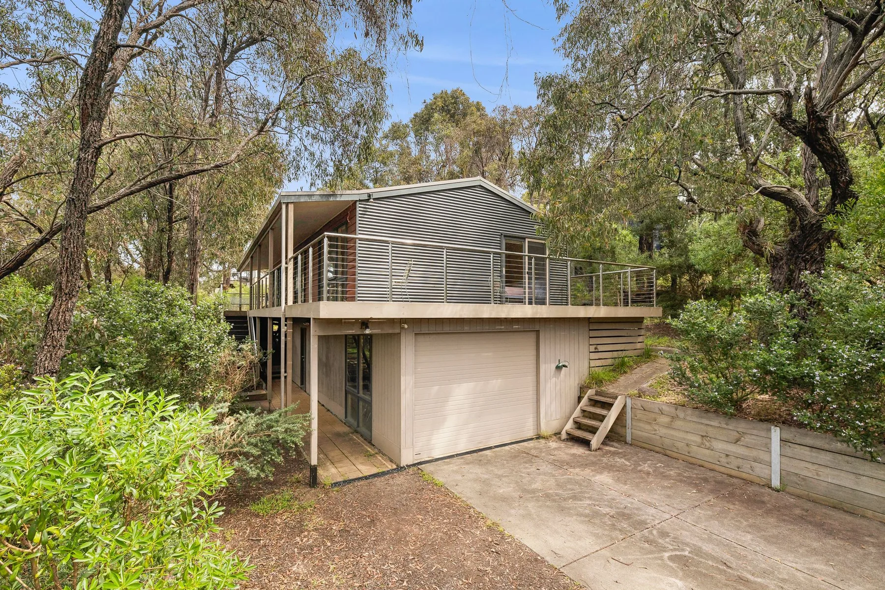 A raised house with a balcony stands among trees beside a paved driveway.