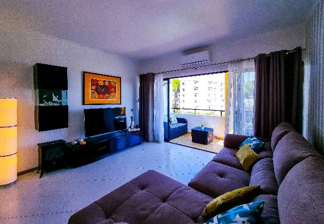 Una sala de estar con un sofá seccional frente a un televisor junto a un balcón abierto.