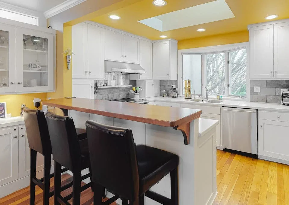 A bright kitchen features white cabinetry, a long counter, and dark seating.