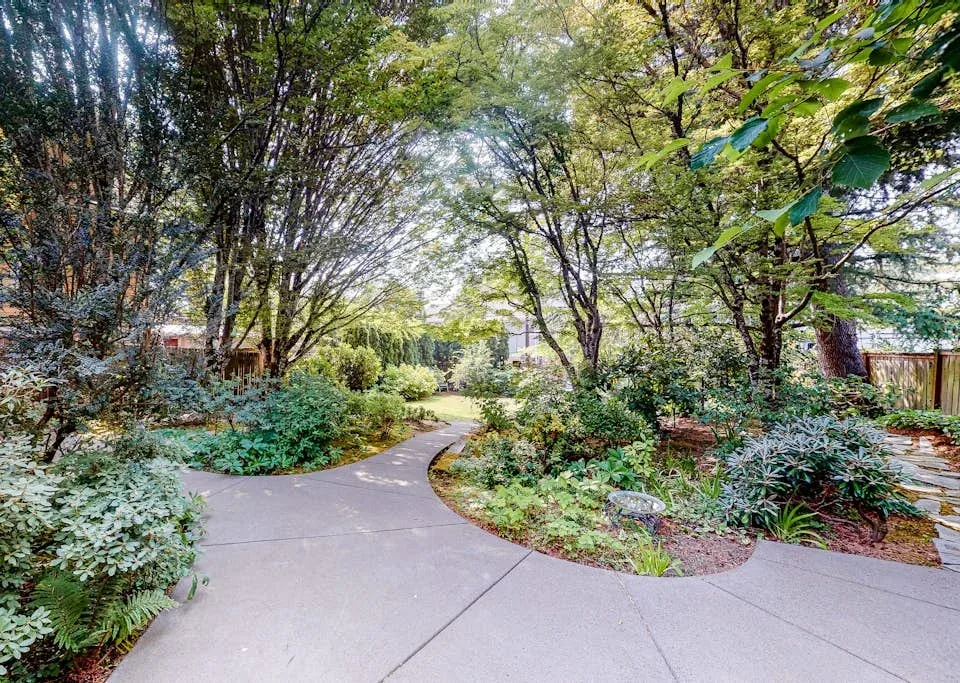 A paved garden path curves through lush trees and planting beds.
