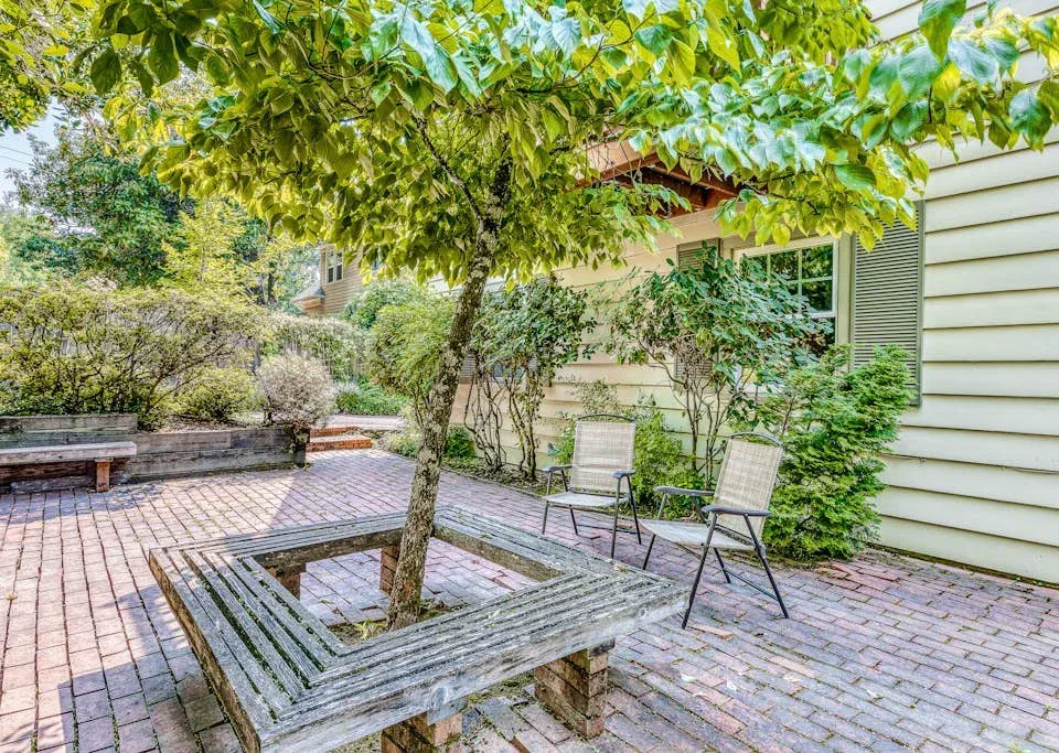 A paved outdoor seating area beneath leafy trees, beside a light-colored house.