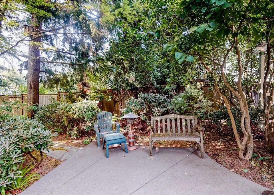 A garden patio with seating sits beneath leafy trees.