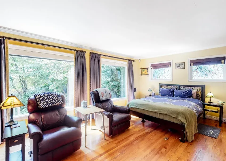 A bedroom with a large bed, two seating areas, and wide windows overlooking trees.