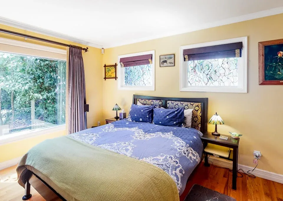 A bedroom with a bed beneath two windows and a large window overlooking greenery.