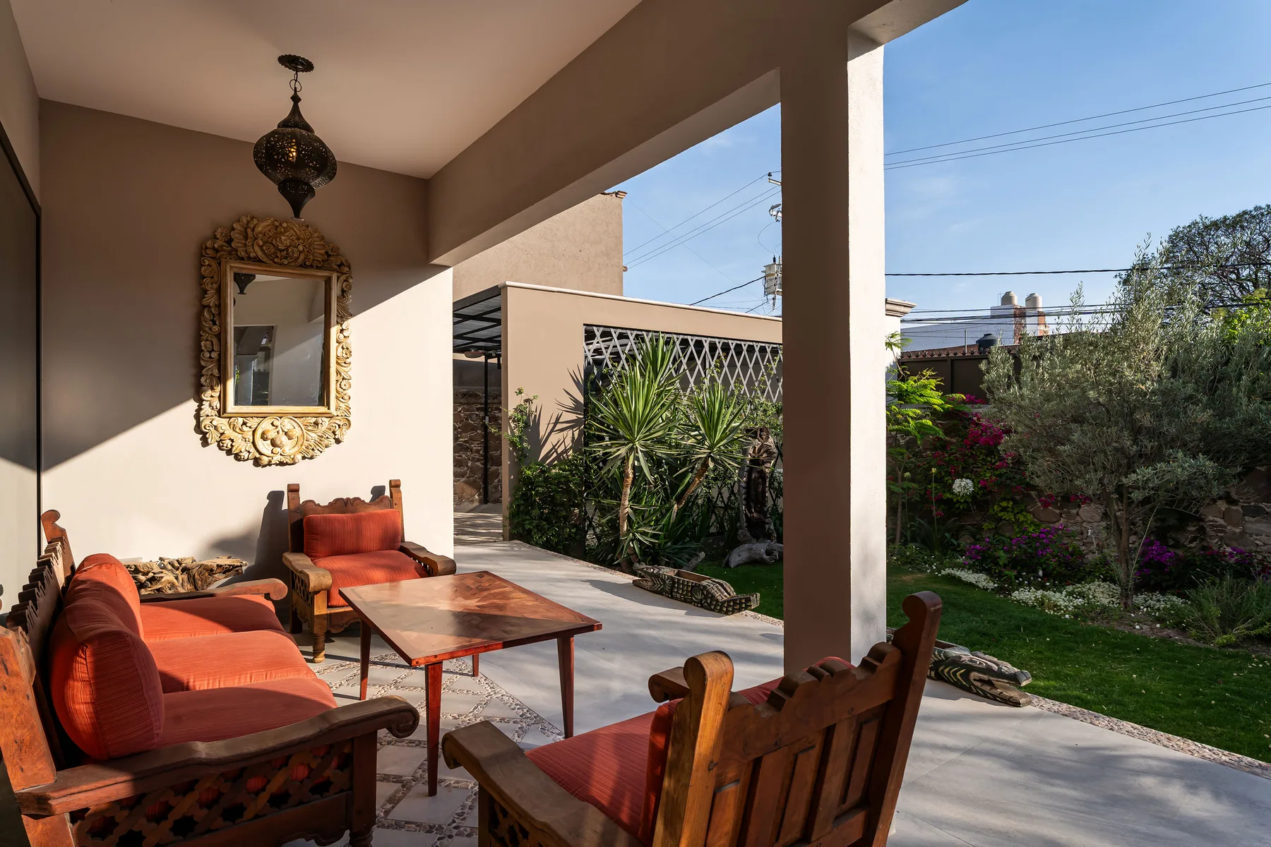 A covered patio has outdoor seating, a low table, and plants along the edge.