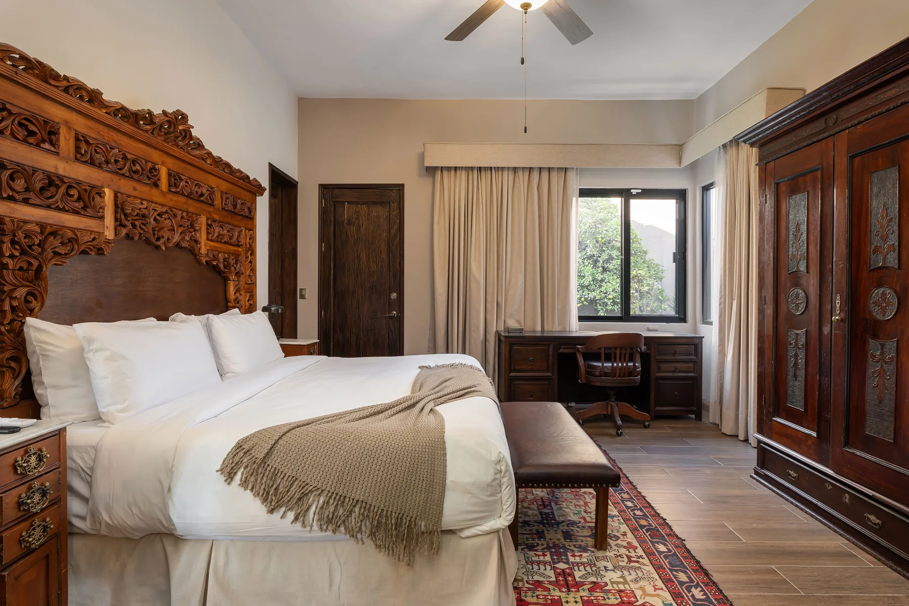 A bedroom with a bed, carved wooden furnishings, and a desk by the window.