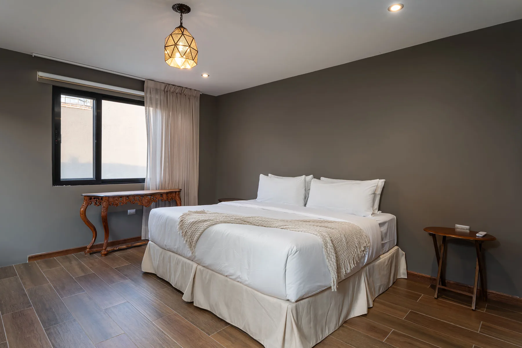 A bedroom with a neatly made bed beside a window and small tables.