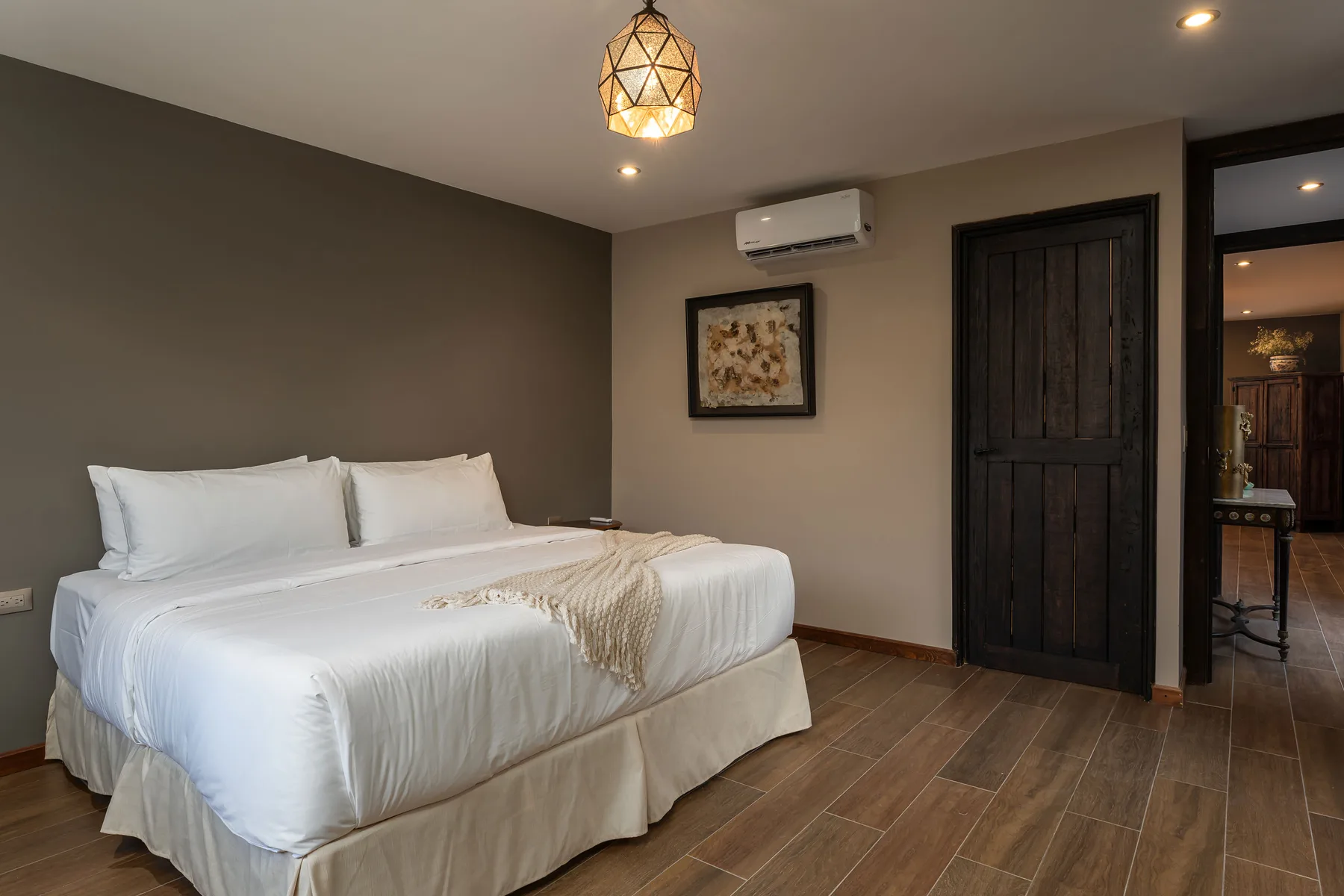 A bedroom with a neatly made bed, framed wall art, and a dark wooden door.