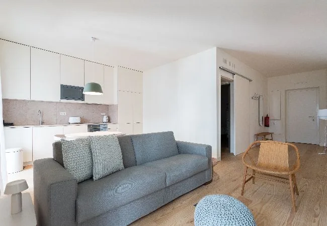 Una luminosa sala de estar diáfana con un sofá gris y la cocina al fondo.