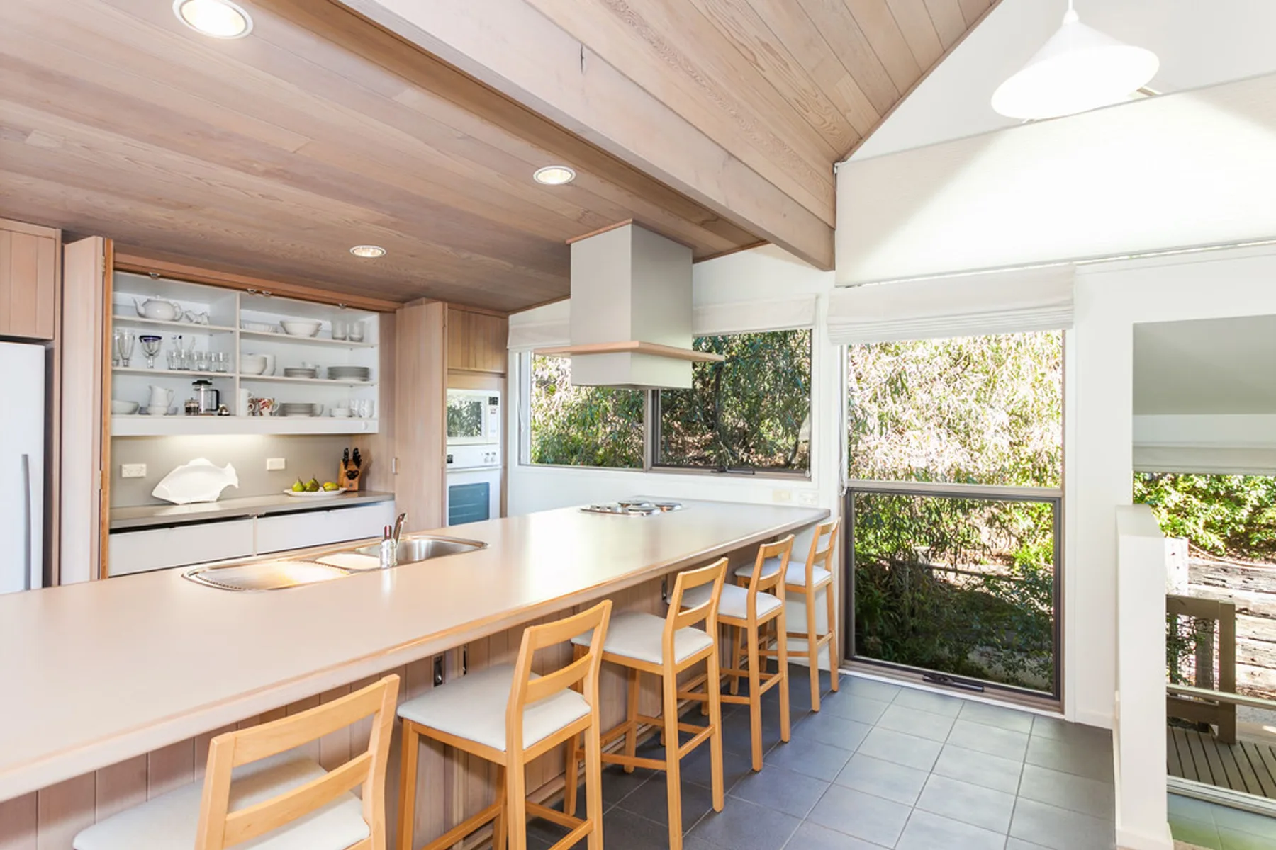 Una cocina luminosa con una isla larga y taburetes de madera junto a grandes ventanas.