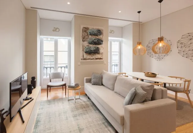 Una sala luminosa con un sofá claro y una mesa de comedor junto a las ventanas.