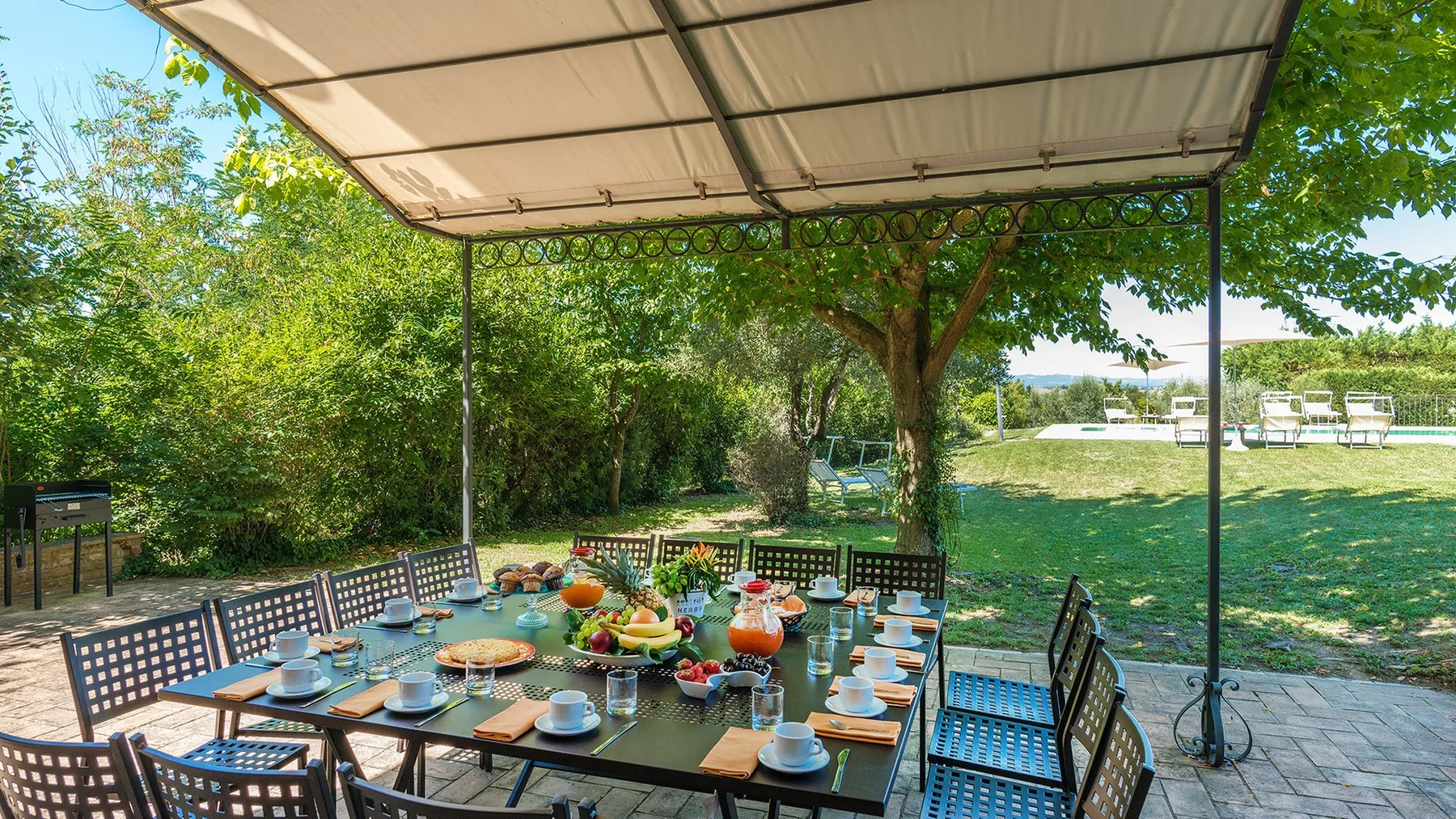 Comedor al aire libre bajo un toldo, con una mesa puesta entre árboles frondosos.