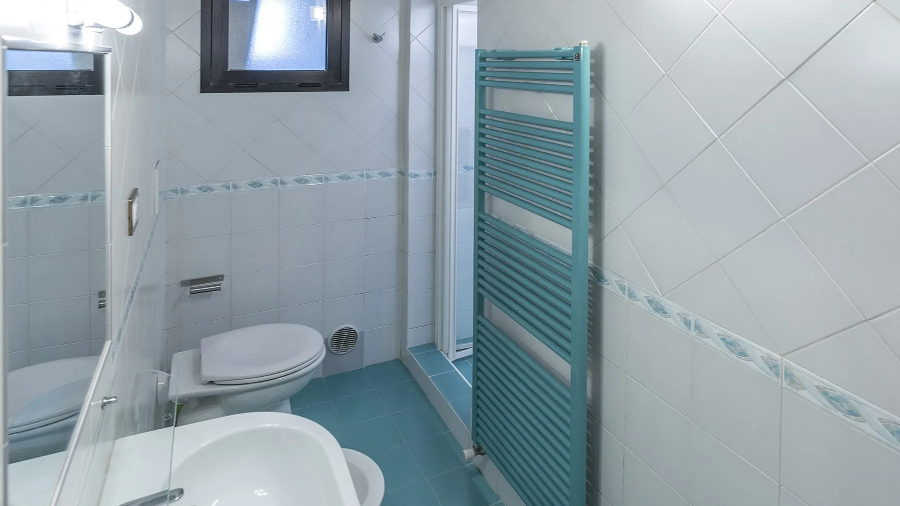 Un baño alicatado con inodoro, lavabo y radiador toallero turquesa.