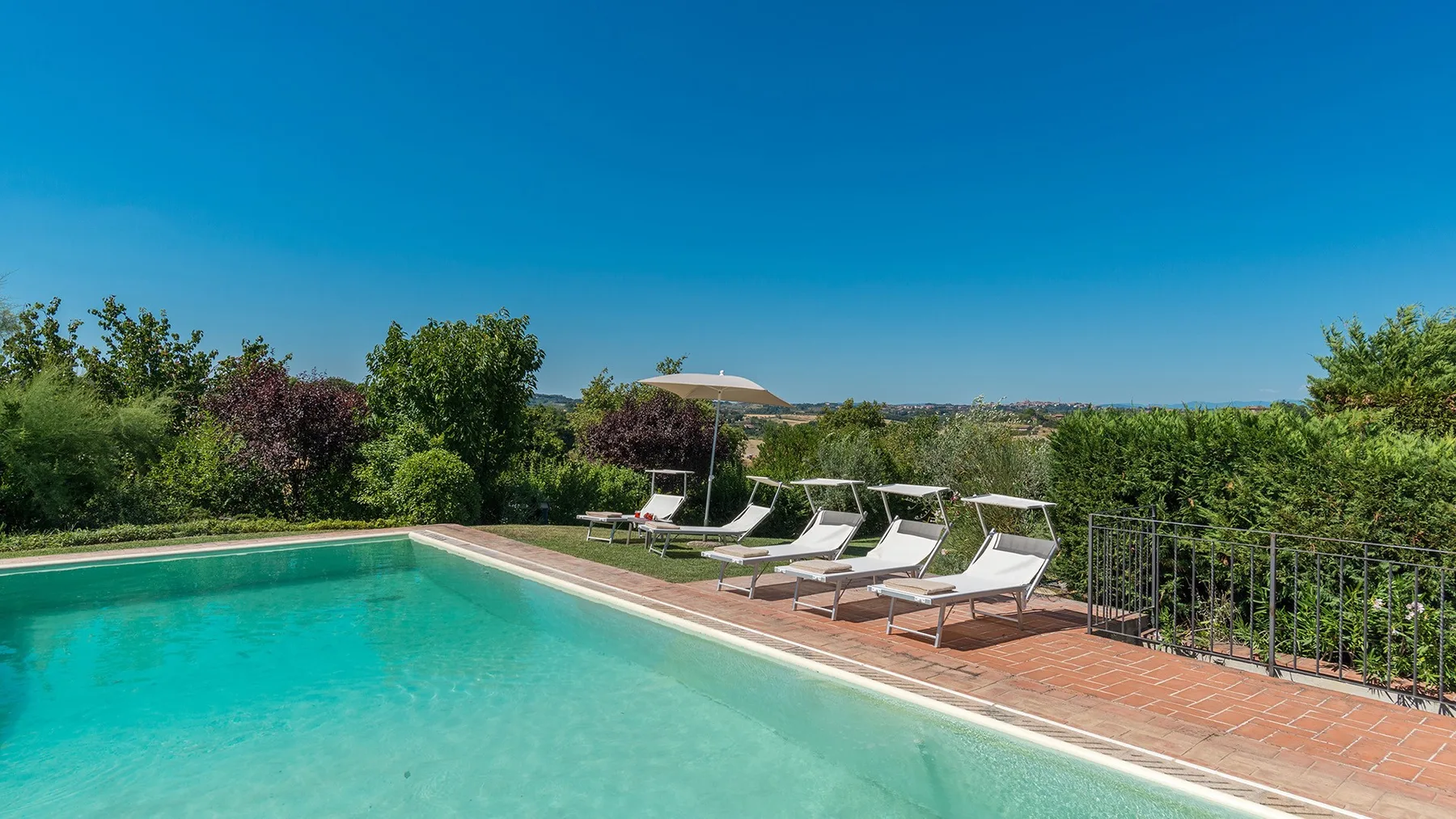 Una piscina exterior junto a una terraza de baldosas, asientos reclinables y vegetación.