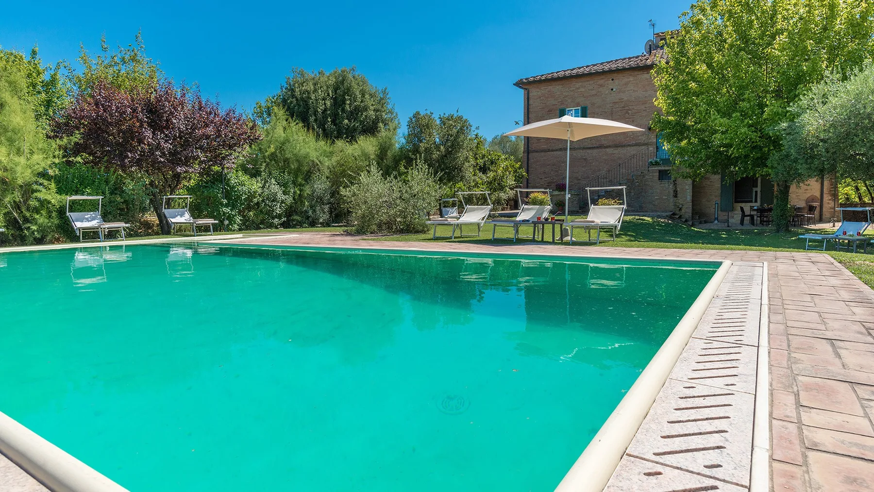 Una piscina junto a una terraza pavimentada, asientos de jardín y un patio sombreado.