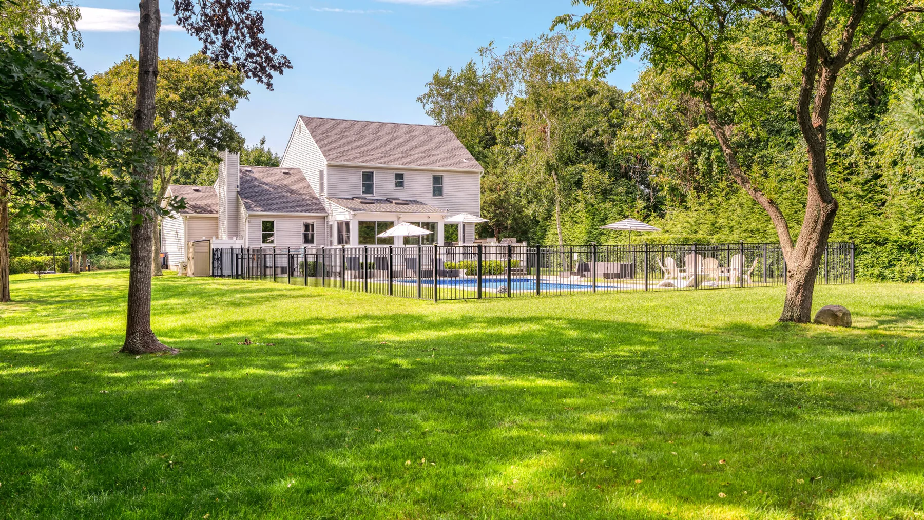 A house and fenced swimming pool sit in a grassy yard surrounded by trees.