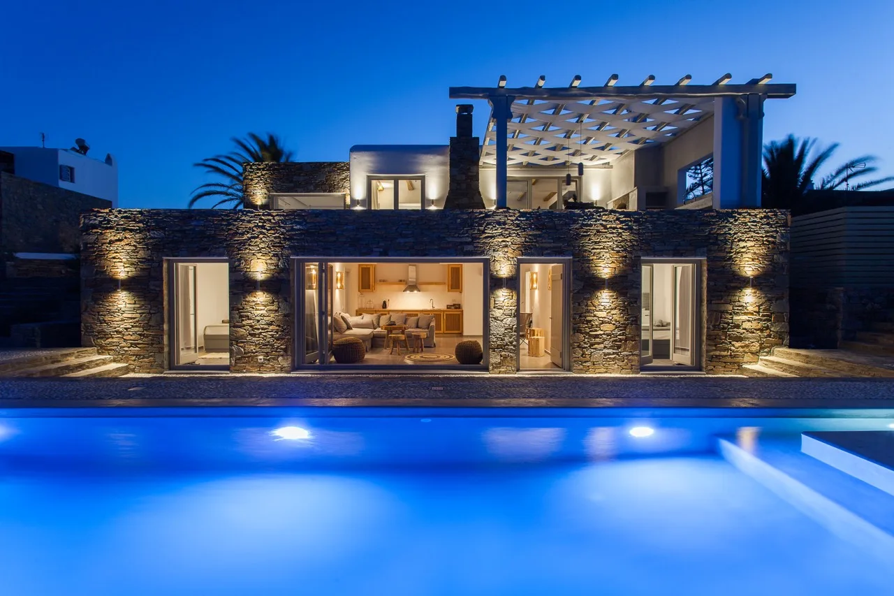 A modern house with a stone façade overlooks an illuminated swimming pool at dusk.