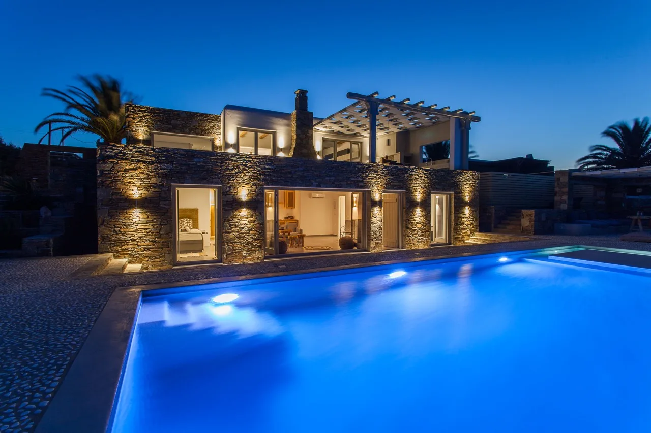 A modern house glows beside a blue-lit swimming pool at dusk.