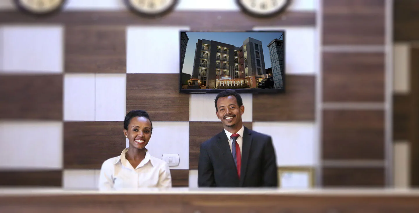 Espacio representativo del hotel: Dos empleados del hotel están detrás de un mostrador de recepción bajo una pantalla.
