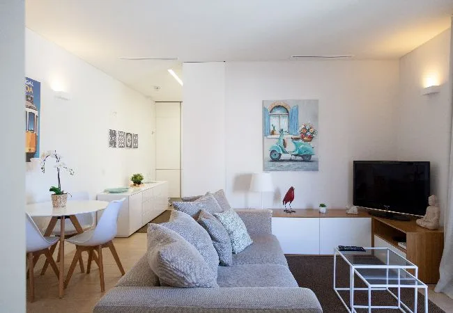 Una sala luminosa con un sofá gris, una mesa de comedor y un televisor.