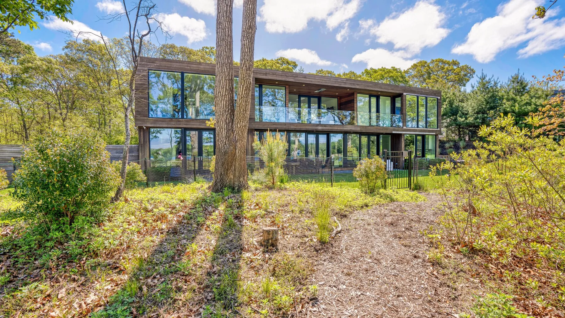 A modern glass-fronted house stands among trees beside a gravel path.