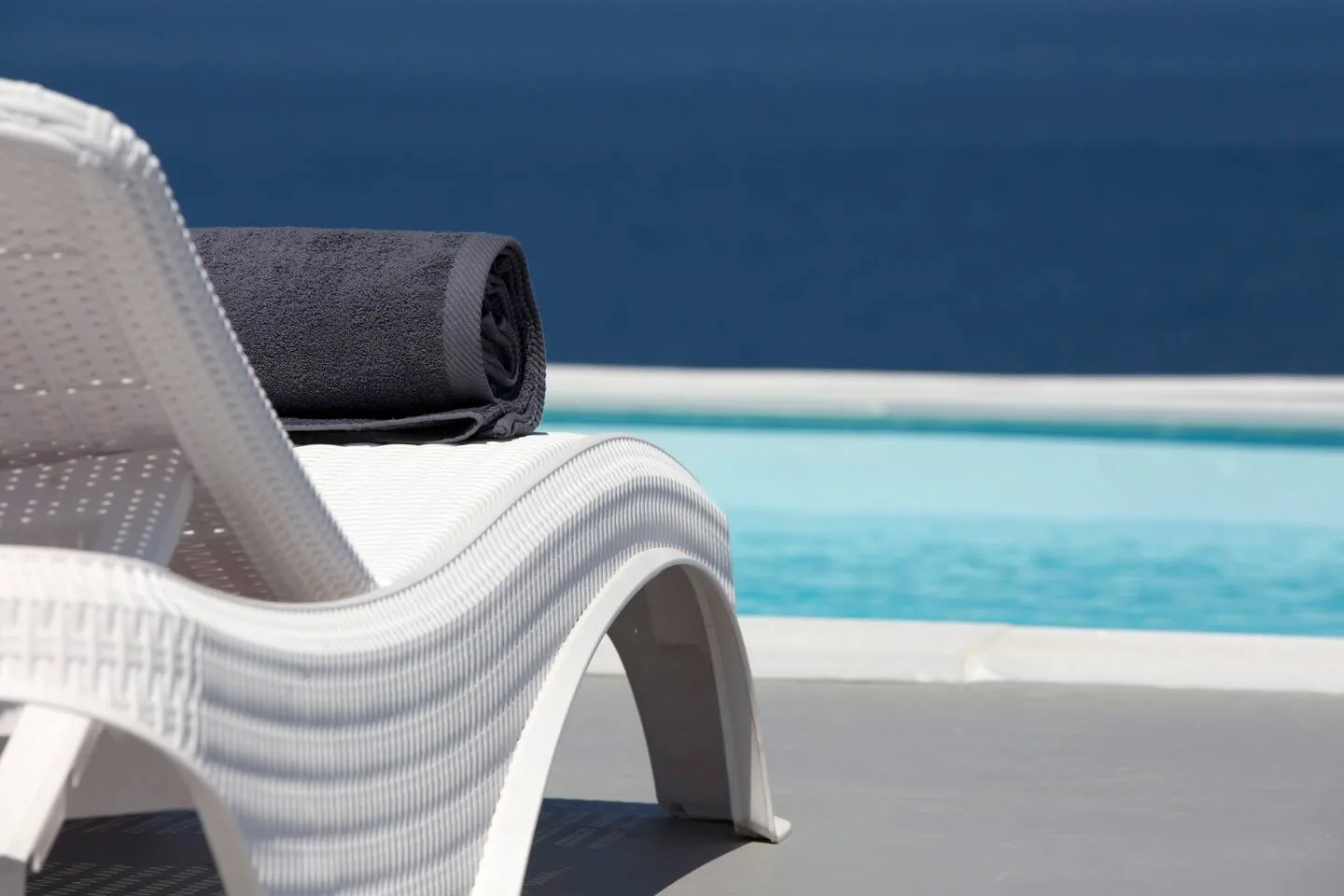 A white lounge chair with a rolled towel sits beside a blue swimming pool.