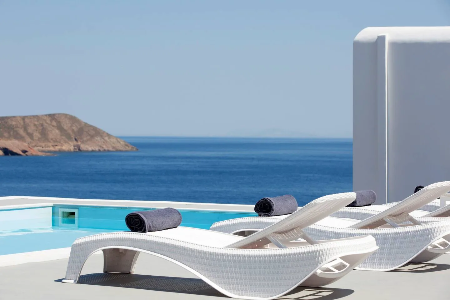 White lounge seating beside a pool overlooks the sea and rocky coastline.