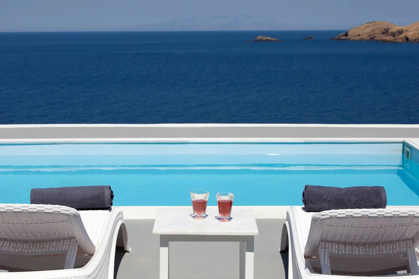 A pool overlooks the blue sea, with lounge seating beside the water.