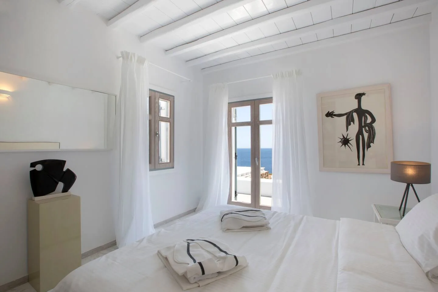 A bright white bedroom with a bed facing tall windows overlooking the sea.
