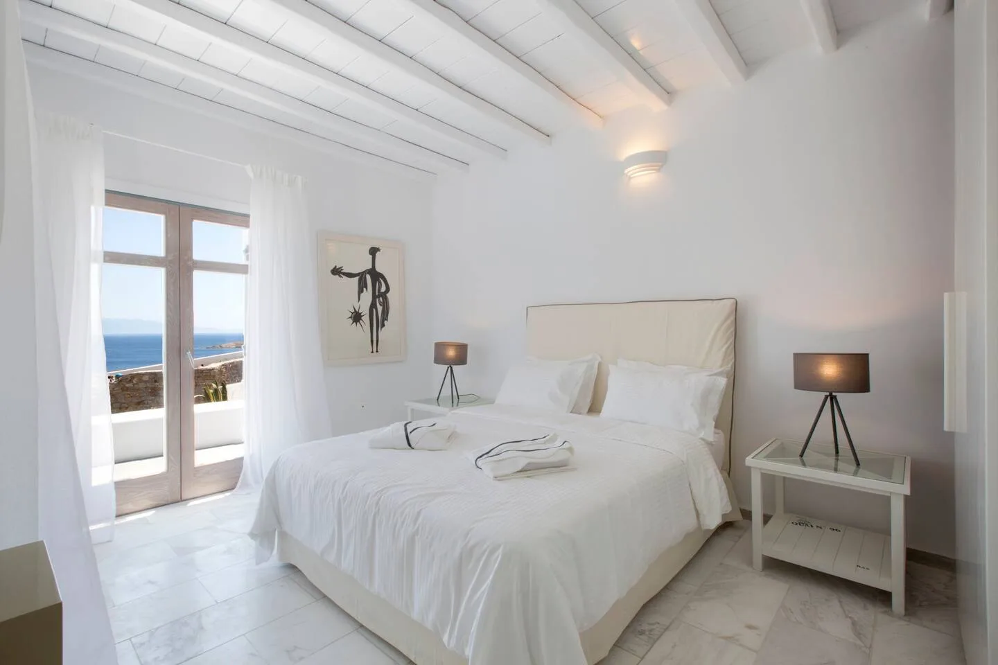 A bright bedroom with a white bed and glass doors opening onto a sea view.