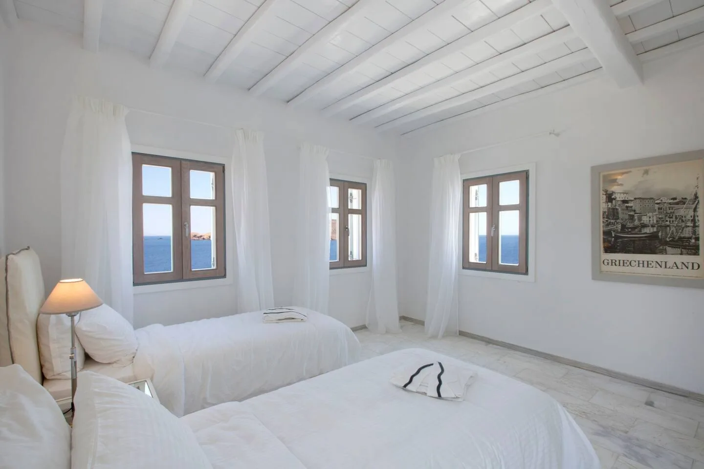 A bright white bedroom with beds, tall windows, and a view of the sea.