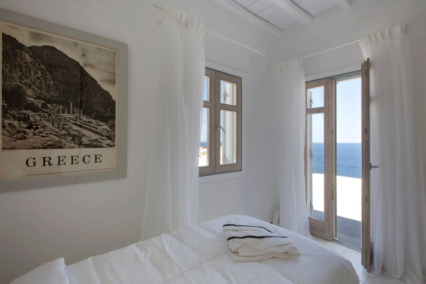 A bright bedroom with a white bed and open doors overlooking the sea.