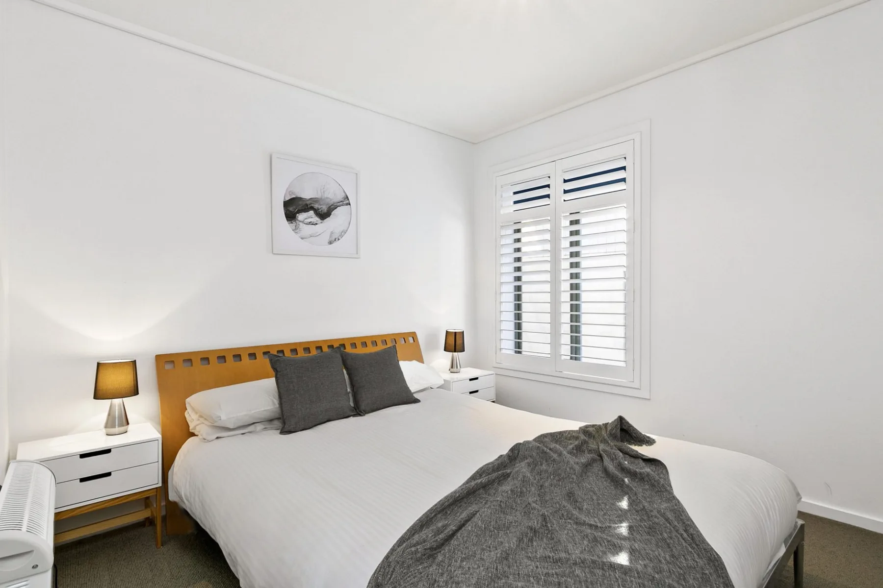 Un dormitorio luminoso con una cama, mesitas de noche y una ventana con persianas.