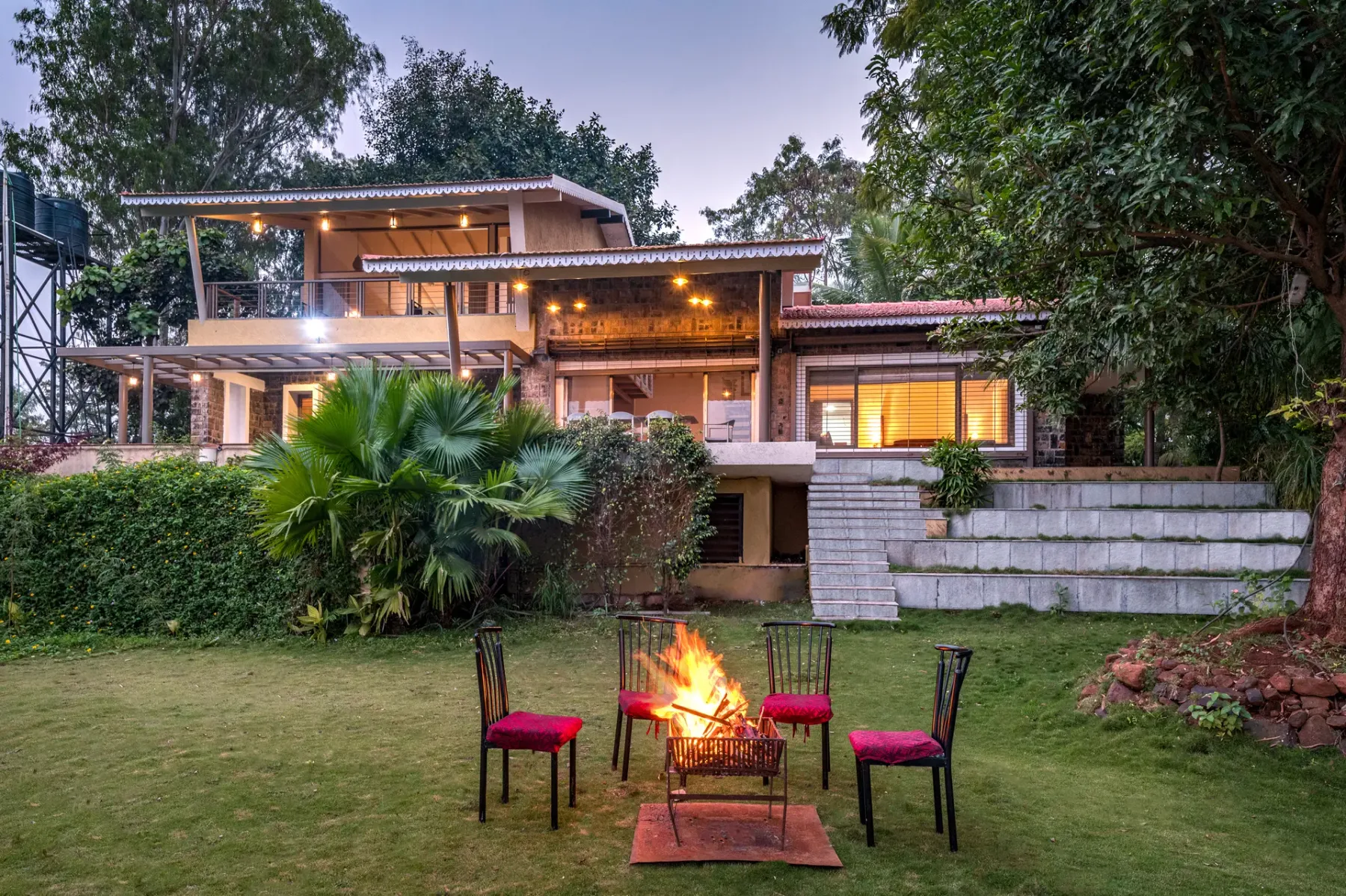 A modern house with a lit patio overlooks a lawn with chairs around a fire.