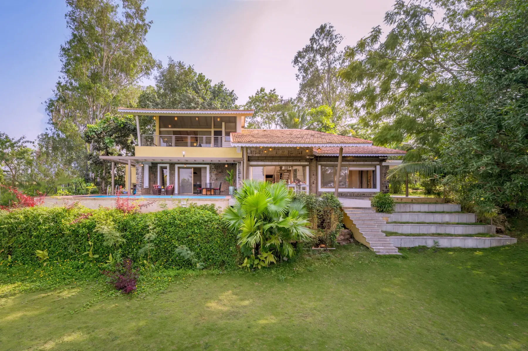 A two-story house sits behind a lawn and landscaped garden.