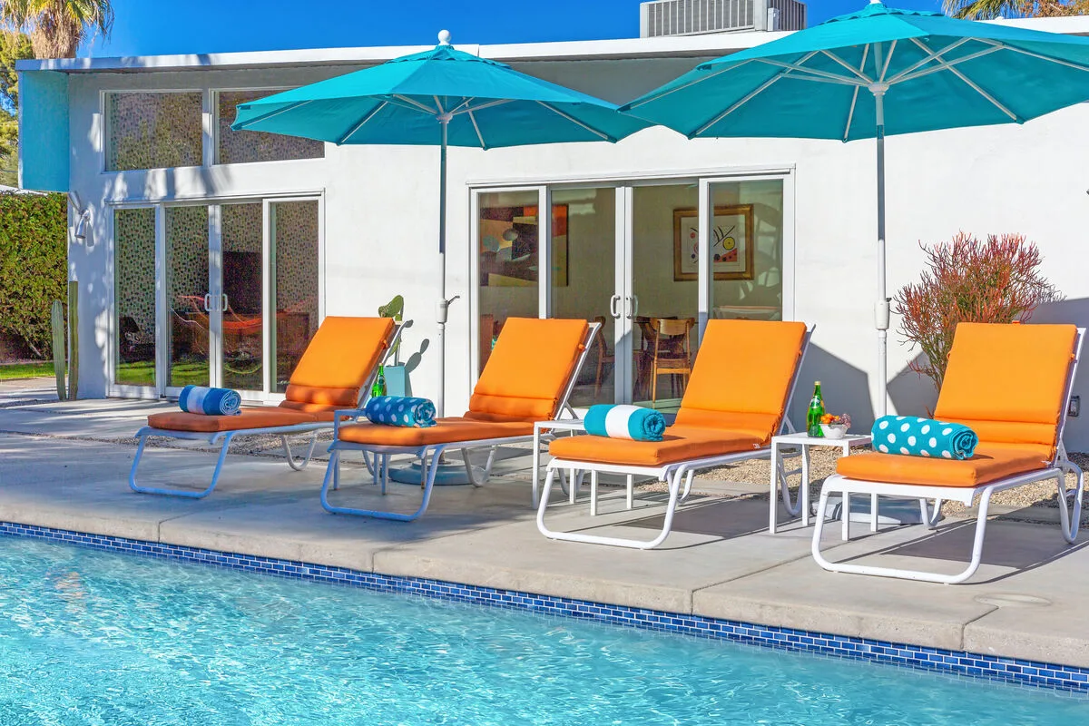 Un patio junto a la piscina tiene asientos naranjas bajo sombrillas turquesas.