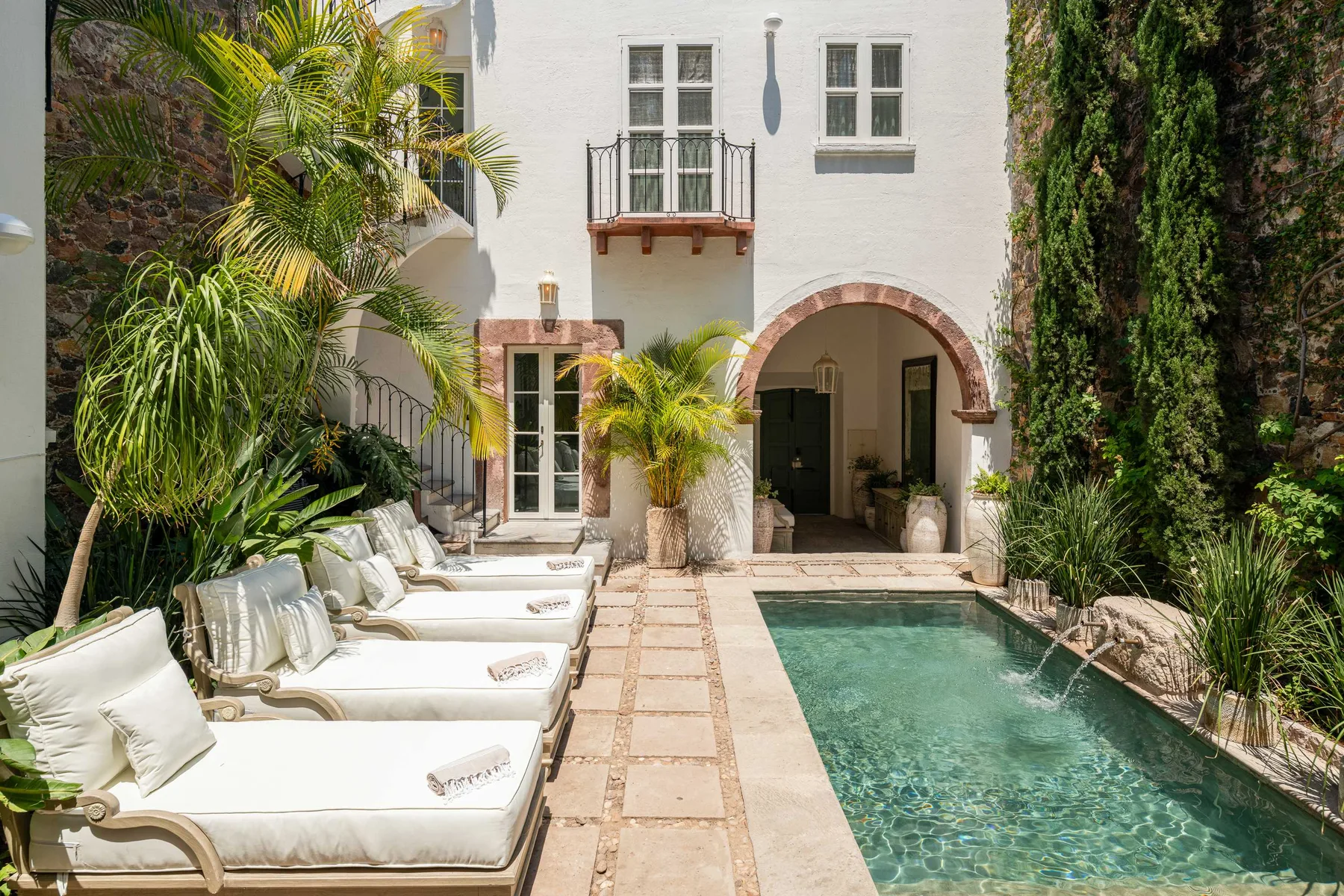 Una piscina de patio está junto a asientos acolchados y un edificio blanco con un arco.