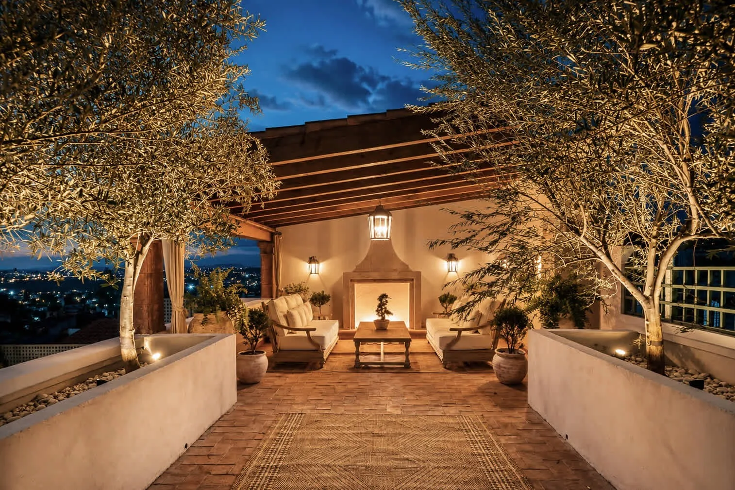 Una terraza exterior iluminada y con asientos bajo una pérgola de madera, enmarcada por árboles.