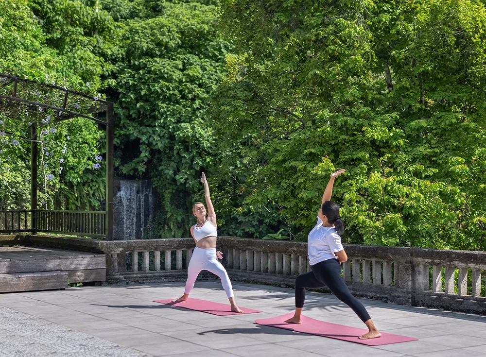 Espacio representativo del hotel: Dos personas se estiran sobre esterillas de yoga en una terraza pavimentada rodeada de vegetación.