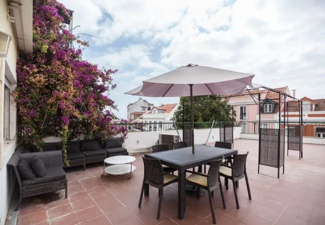 Una terraza exterior con suelo de baldosas, mesa de comedor, asientos y una gran sombrilla.