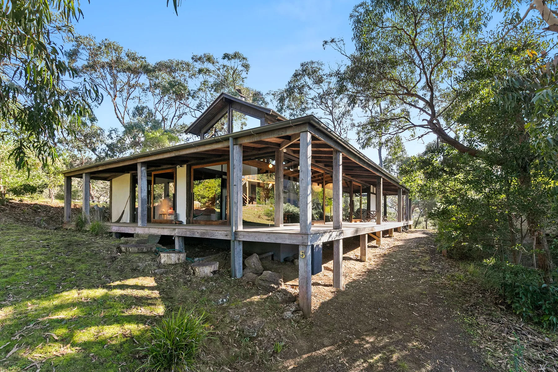 A raised timber house stands among trees on a sloping, grassy site.