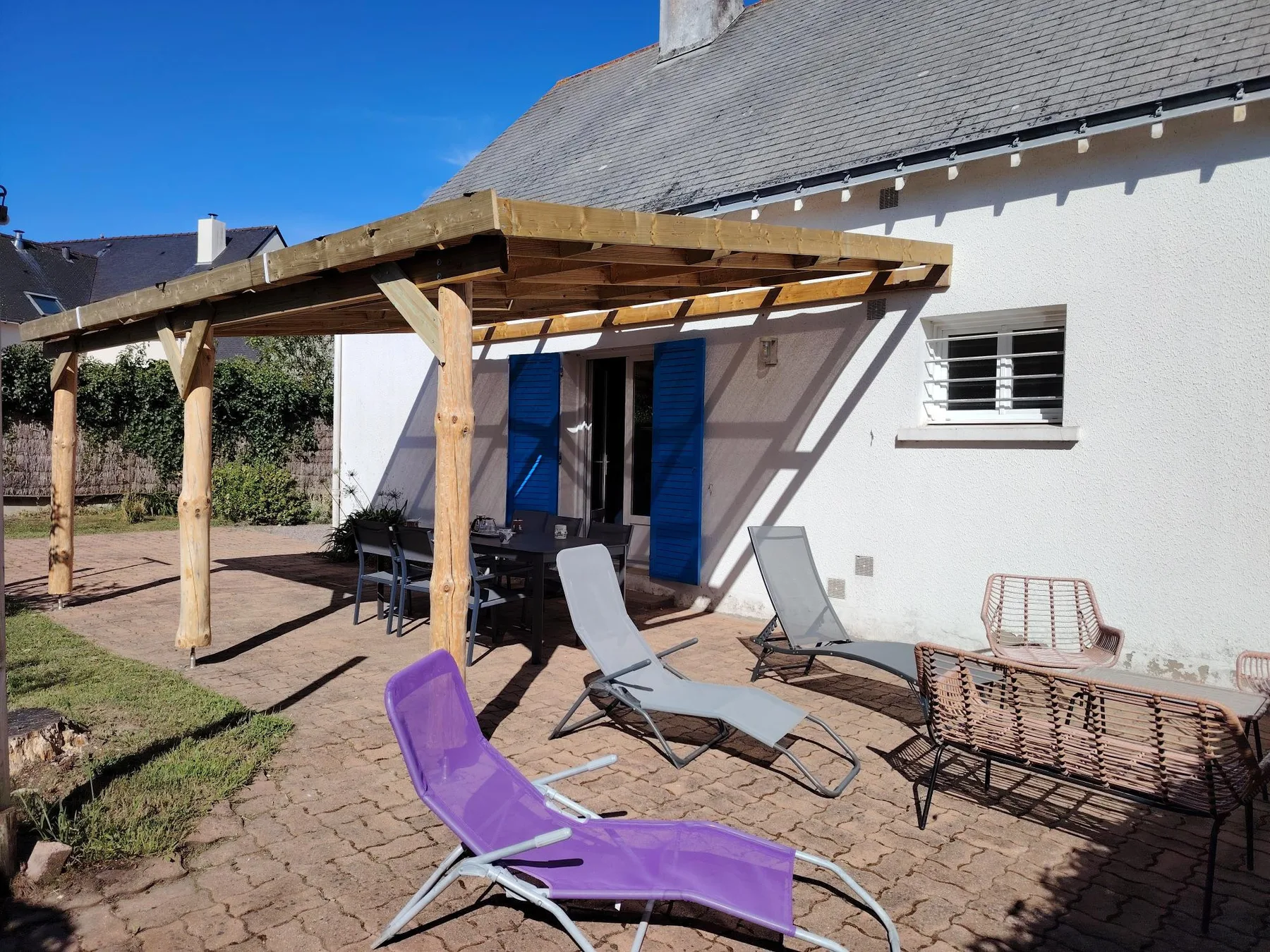 A sunny outdoor patio has lounge seating beneath a wooden pergola.
