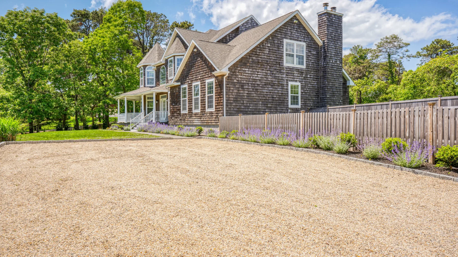 A shingle-sided house stands beside a broad gravel driveway and wooden fence.