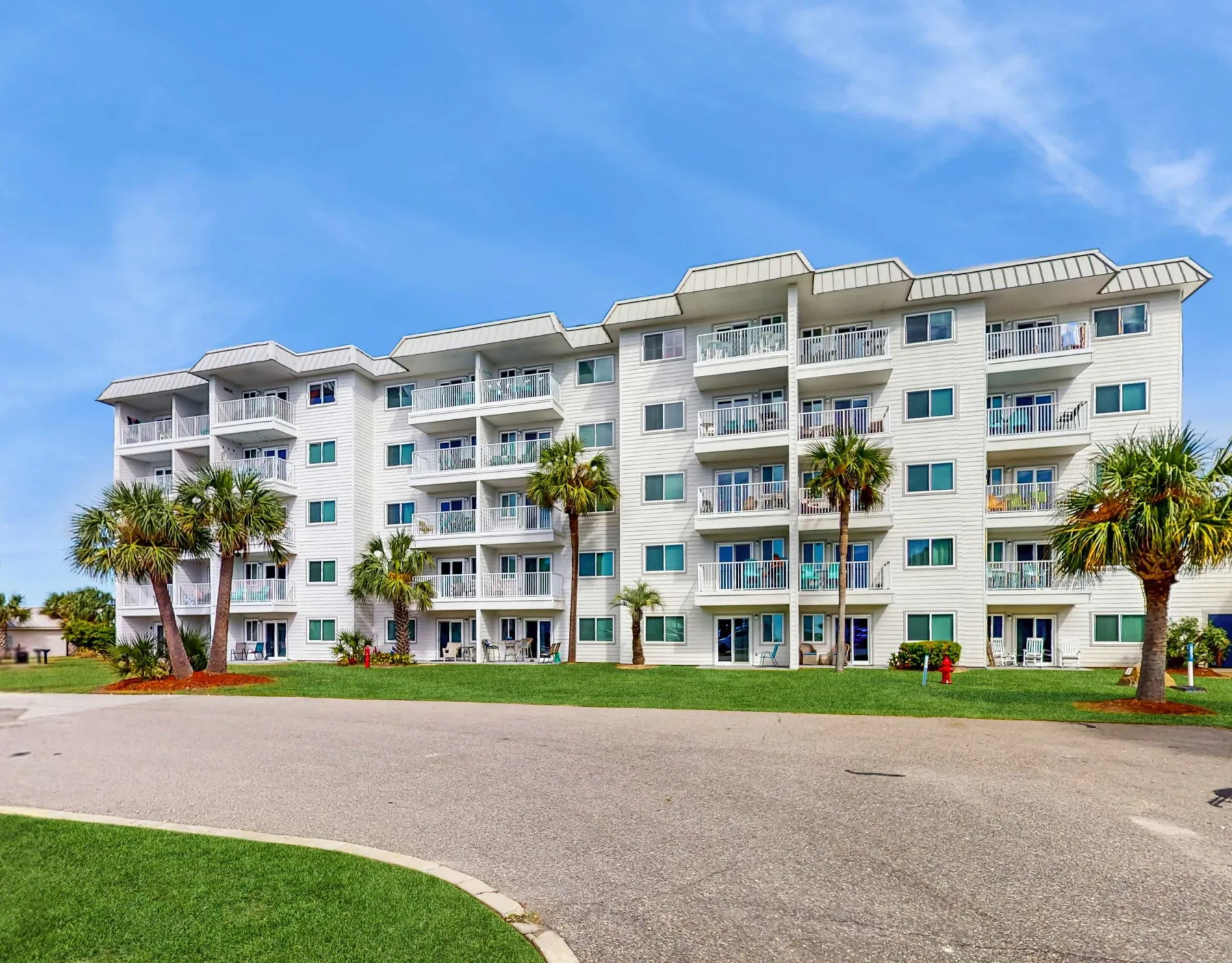 Zona común: Un edificio de apartamentos de varios pisos está junto a una entrada pavimentada y palmeras.
