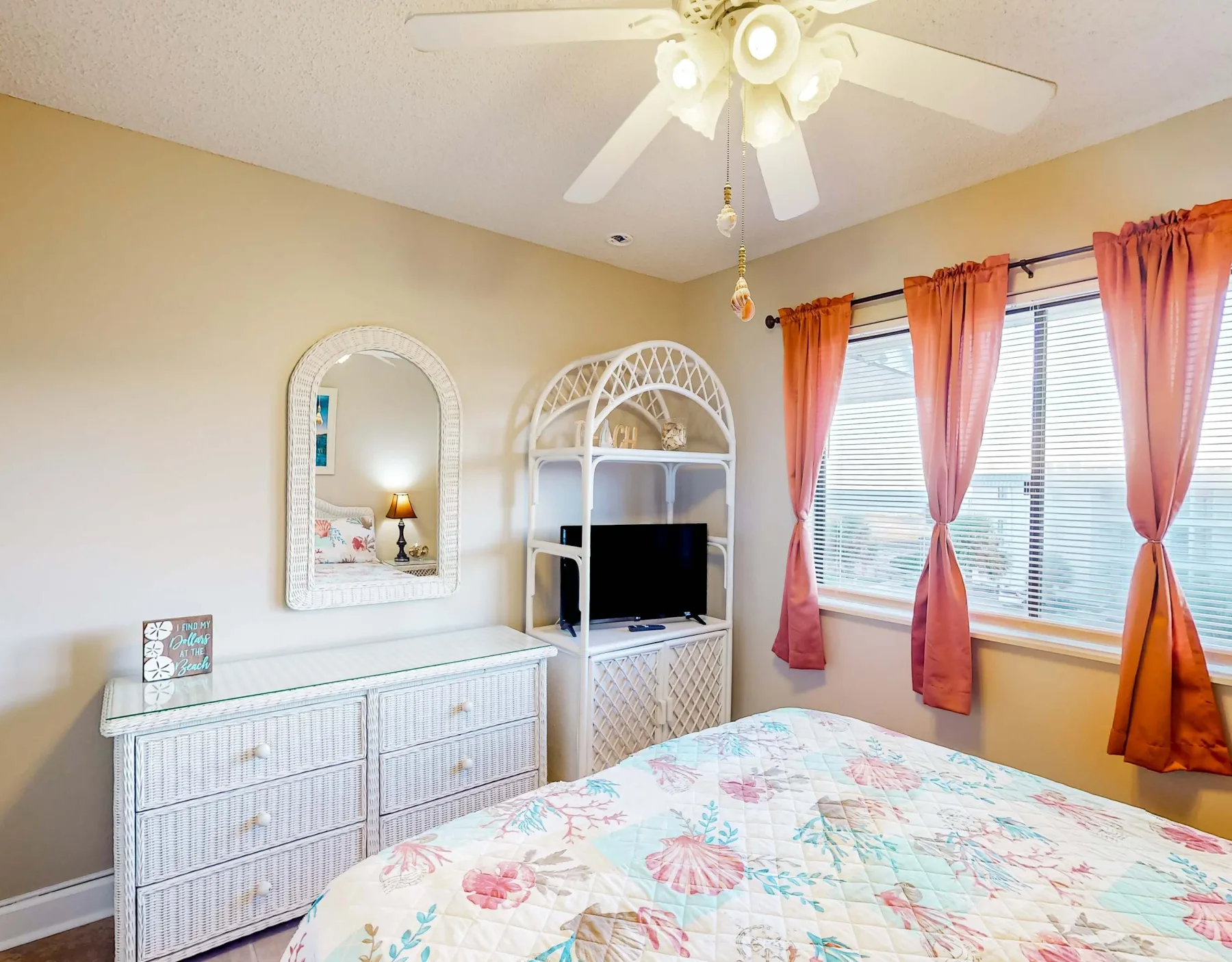 Un dormitorio con una colcha floral, una cómoda blanca y ventanas con cortinas coral.
