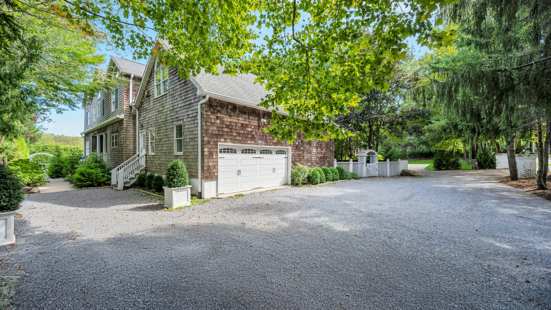 A large house stands beside a broad gravel driveway, surrounded by trees.