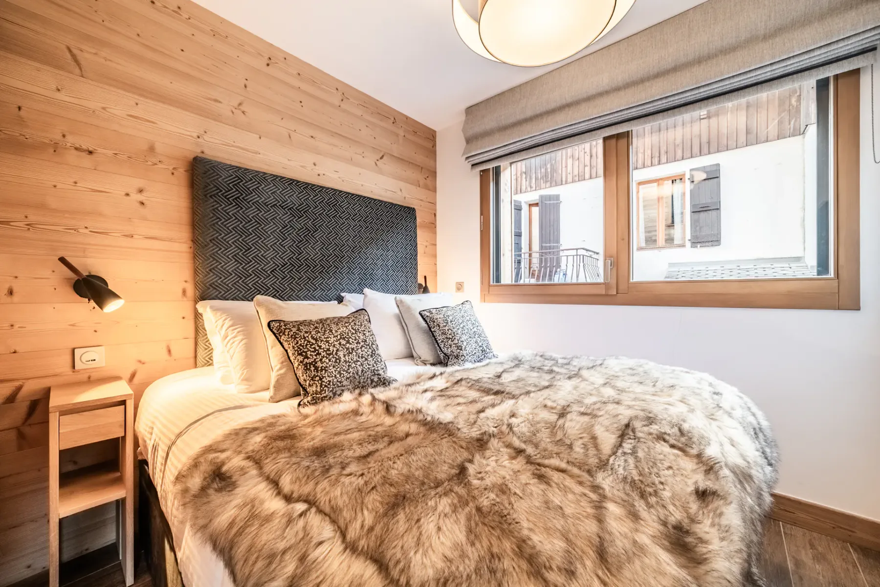 A bedroom with a bed beneath a large window overlooking nearby buildings.