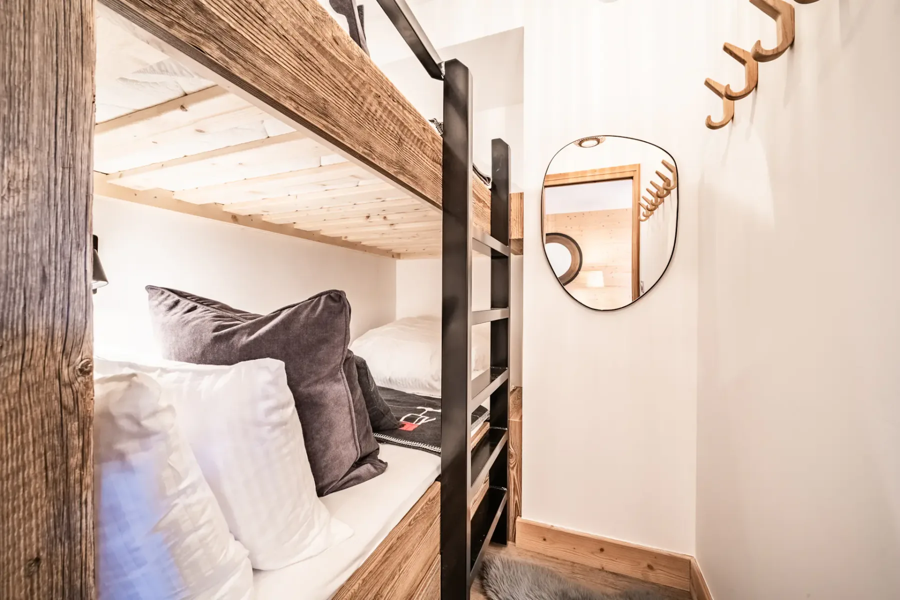A bunk bed with pillows sits beside a wall mirror and coat hooks.