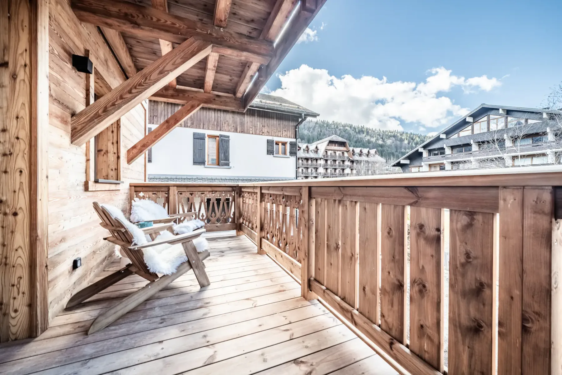 A wooden balcony with seating overlooks nearby buildings and a forested hillside.